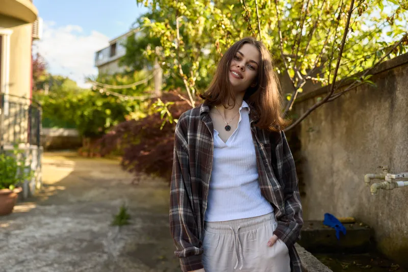 Cheerful woman outdoors, casual lifestyle, wearing a plaid jacket over a white shirt, standing in a sunlit street with greenery, relaxed pose, gentle breeze, warm tones