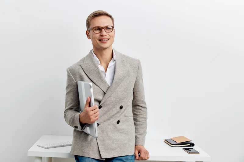 smiling young man holding laptop wearing glasses beige jacket standing in modern office digital nomad concept