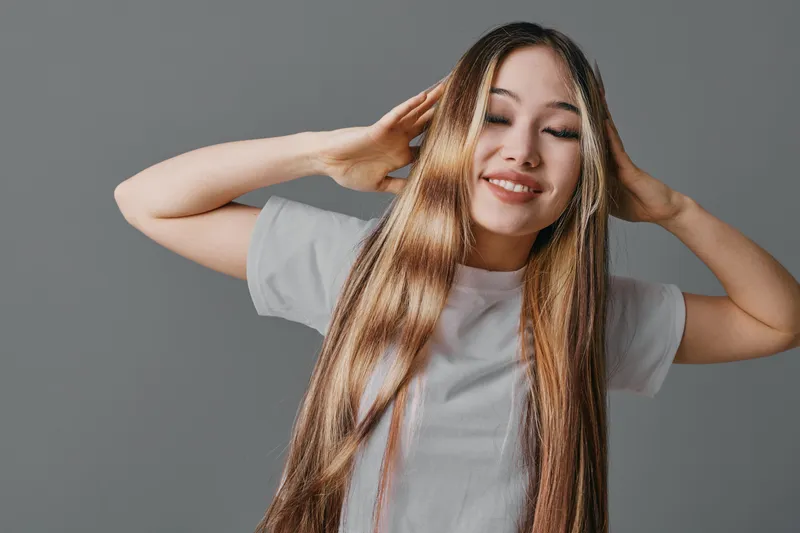 Young woman with long hair posing with closed eyes and hands near head, smiling gently in casual white t shirt on grey background for beauty and relaxation concept.