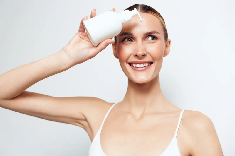 Young woman with clean fresh skin holds white lotion bottle near face smiling happily isolated on light background skincare concept beauty routine moisturizing product.