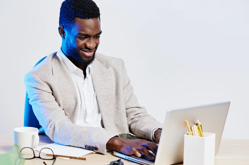 Professional young man working on laptop at desk, smiling and focused, in a modern office environment, casual business attire, workspace with stationery and coffee, concept of remote work and producti