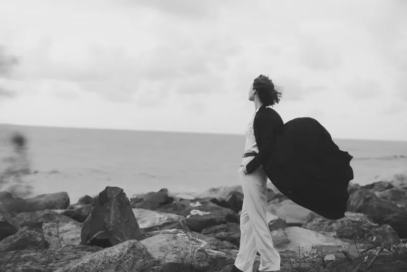 Woman with flowing cape walks along a rocky shore toward the ocean, wind catching fabric, monochrome grayscale landscape conveying solitude, journey and resilience