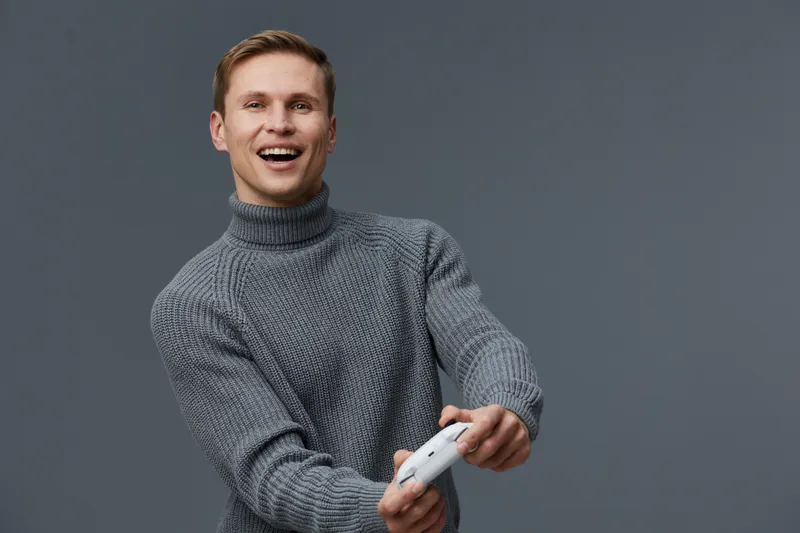 Smiling young caucasian man wearing gray turtleneck sweater playing video game controller on plain gray background casual lifestyle concept