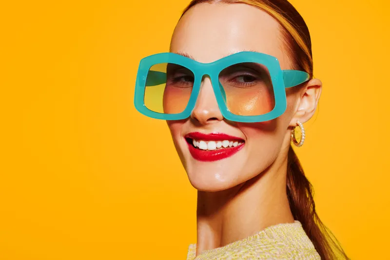 Smiling woman with large turquoise glasses and red lipstick poses against bright yellow background. Fashion portrait with vibrant colors and confident expression. Concept of style and beauty.