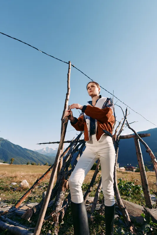 Woman standing by rustic fence in mountains, wearing tall boots and stylish jacket, outdoor fashion portrait with clear blue sky, natural landscape and confident strong pose.