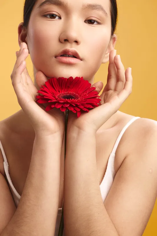 Asian woman holds a red flower close to her face against a warm yellow backdrop, creating a soft, artistic portrait with calm expression and graceful poise.
