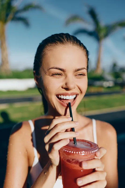 A young woman in a white tank top smiles while sipping a pink smoothie in a sunlit outdoor setting, conveying a casual lifestyle moment with relaxed posture and a vibrant, tropical backdrop.