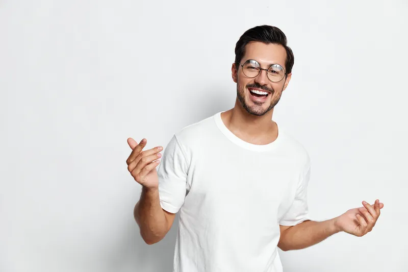 Smiling young man wearing glasses and a white t-shirt, joyfully gesturing with hands against a plain light background. People lifestyle concept.