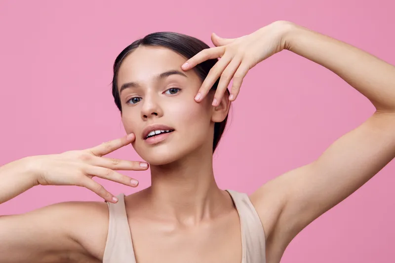 Young woman of mixed ethnicity posing gracefully against a pink backdrop, showcasing natural beauty and a fresh, glowing look with hands elegantly framing her face