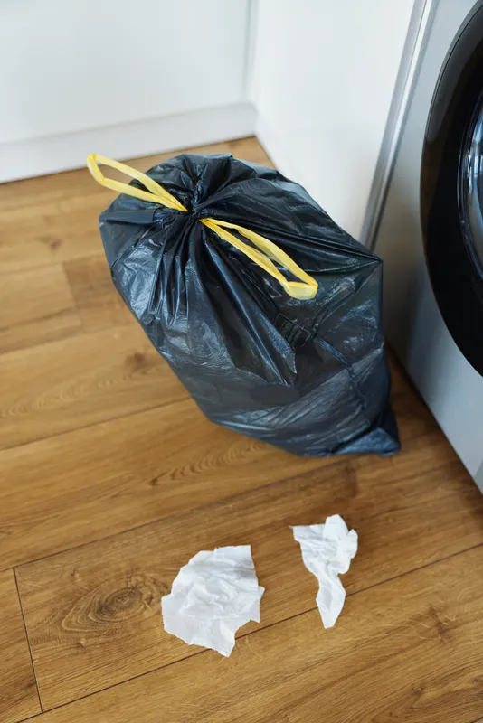 Trash garbage bag waste in laundry room, plastic bag tied with yellow tie beside washer on wooden floor with crumpled tissues, household cleaning and disposal scene for domestic chores.