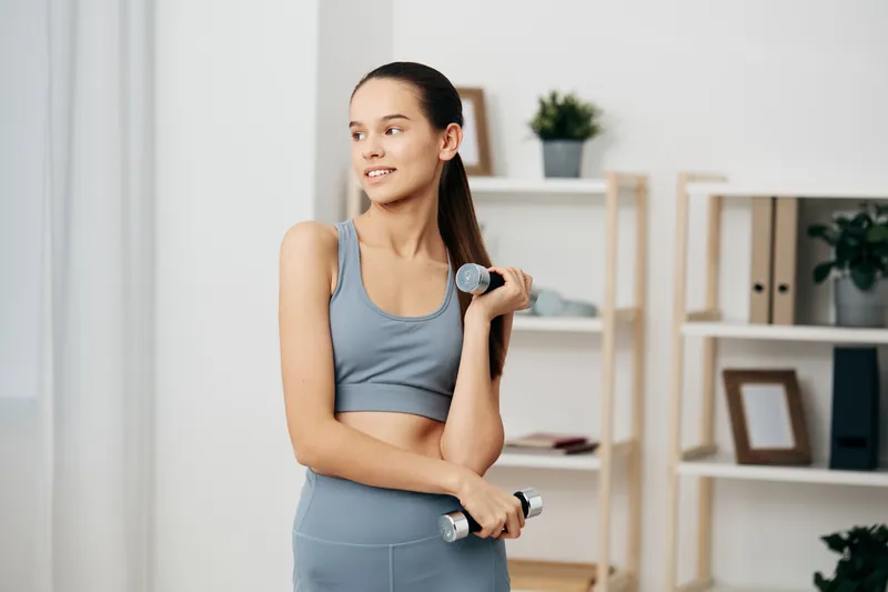 Fit young woman in matching gray sports bra and leggings lifting small dumbbells in a bright home workout setting, confident posture, ponytail, minimalist shelving and plants in soft focus.