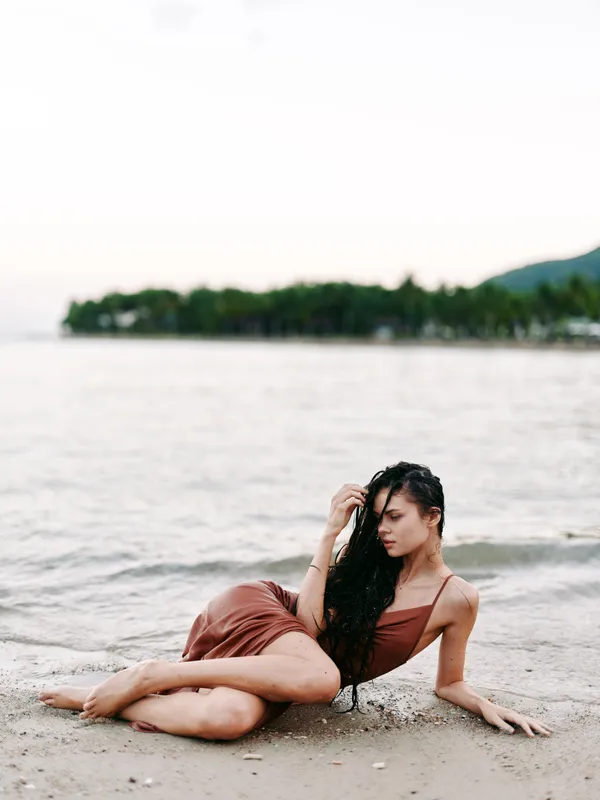 Elegant beach portrait featuring a woman in a brown swimsuit lounging on sandy shore, with distant trees and gentle waves creating a serene summer scene.