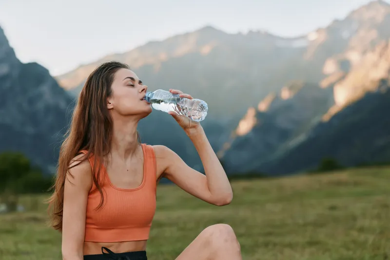 A young woman sits on the grass in a mountain landscape, drinking water from a bottle after a workout, wearing an orange sports top and enjoying fresh air and sunshine.