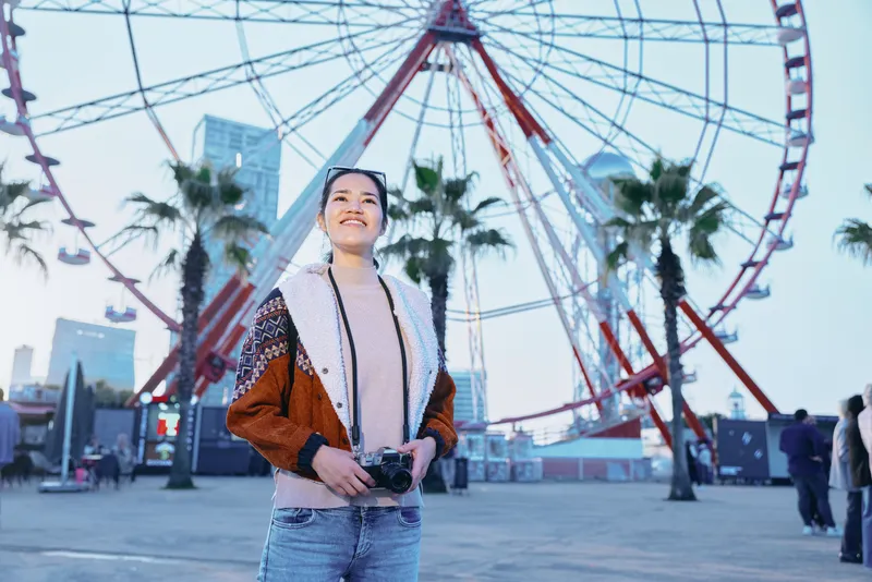 Woman with camera at an outdoor amusement park near a tall ferris wheel, palm trees and city skyline, capturing travel photography memories on a sunny day as visitors stroll by