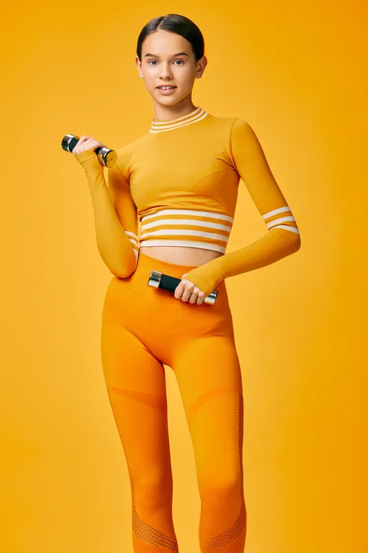 Female athlete in bright orange activewear holding dumbbells against a vivid orange backdrop. Dynamic fitness scene highlighting strength, training and energy for sport or wellness campaigns.