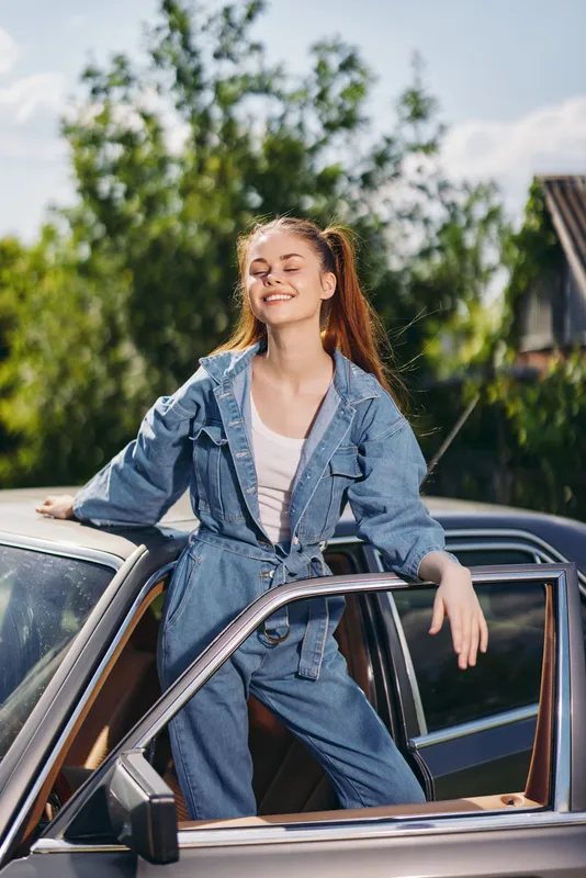 Young woman in denim jumpsuit enjoying a sunny day, posing on a classic car with a bright smile, showcasing joy and carefree emotions in a vibrant outdoor setting.