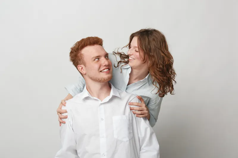 Happy young couple in white shirts smiling and embracing each other on light background. Concept of love, friendship, relationship, and positive emotions between man and woman.