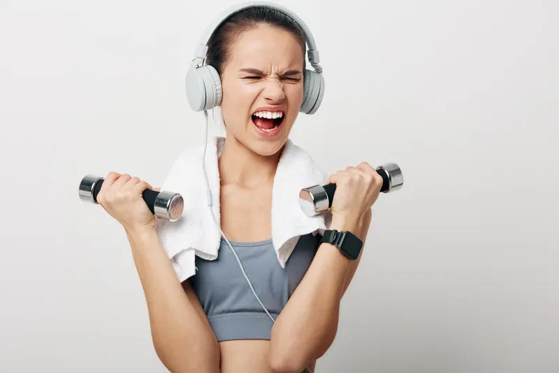 Female fitness enthusiast lifting dumbbells with headphones and a towel around her neck, showcasing strength, focus, and determination during a high-energy workout session in a clean studio.