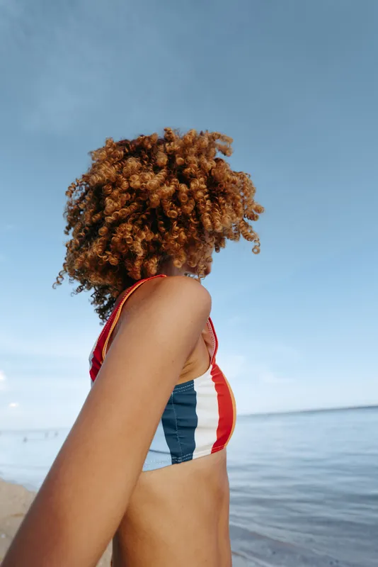 Woman with curly hair in a striped swimsuit stands on a beach near the sea, blue sky behind, side view, outdoors, calm summer mood, soft sunlight and breeze, relaxed vacation portrait in peace