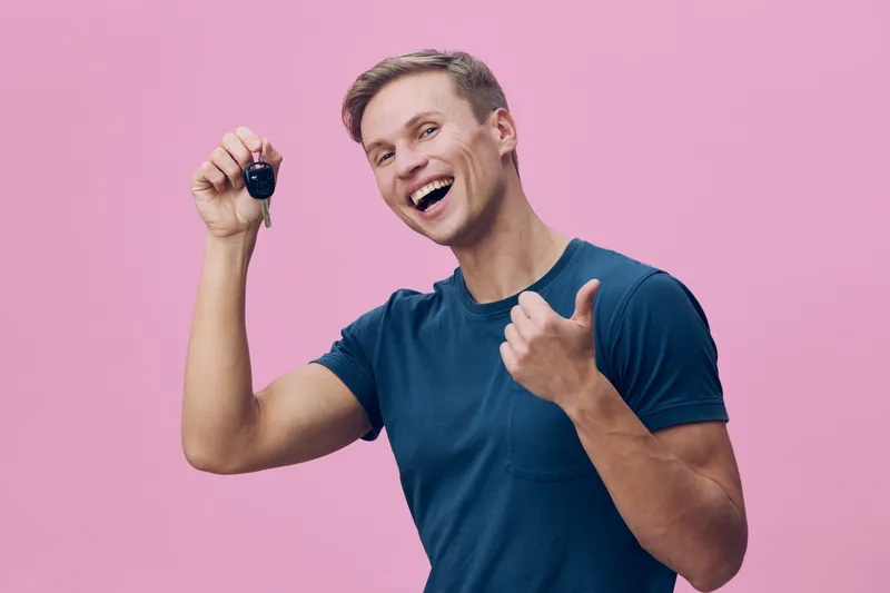 Smiling man in casual blue t-shirt holding car key and giving thumbs up, cheerful expression, positive mood, studio shot with pink background, lifestyle concept, happy and confident.