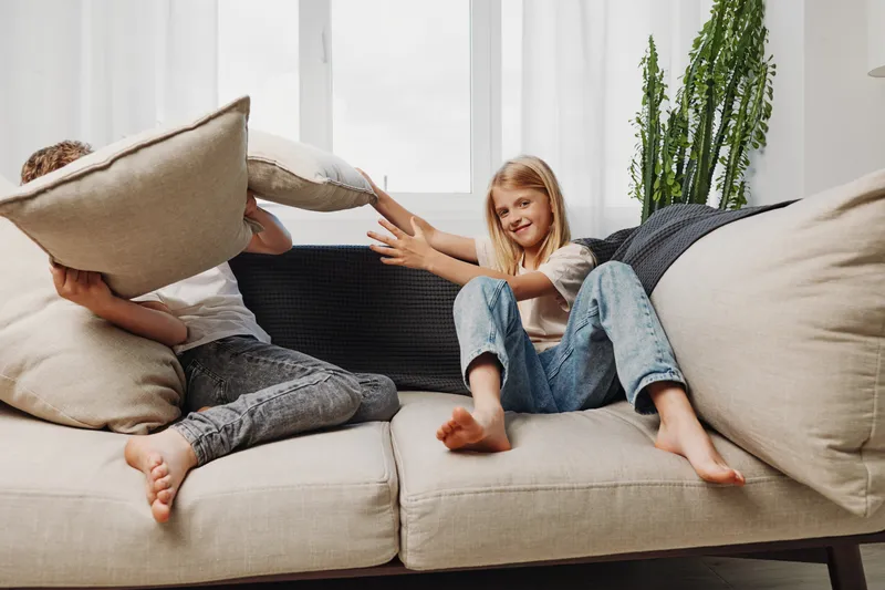 Two children playfully fight with pillows on a beige sofa in a bright living room. The girl smiles while the boy hides behind a cushion in a joyful home setting.