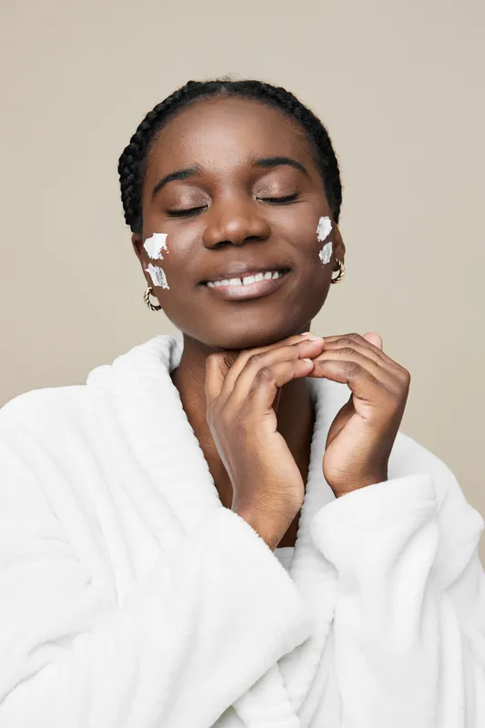 Young Black woman enjoying skincare routine with cream on her face, wearing a cozy robe, and a happy smile against a soft neutral background