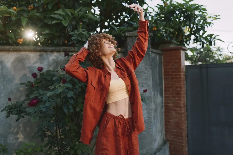 A woman in a rust red outfit stretches arms toward the sky in a sunny outdoor garden, conveying carefree lifestyle, confidence, and contemporary fashion