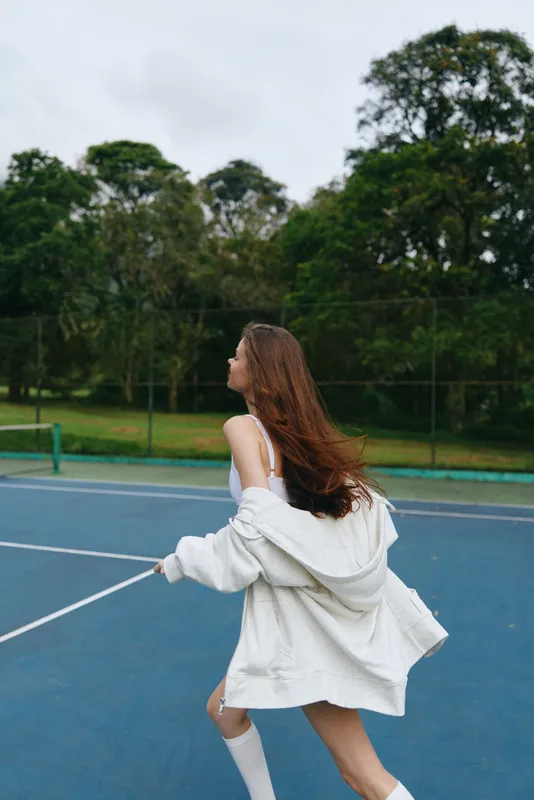 Young woman in a white oversized sweater playing tennis outdoors, expressing joy and freedom, surrounded by lush greenery, capturing a vibrant and sporty atmosphere