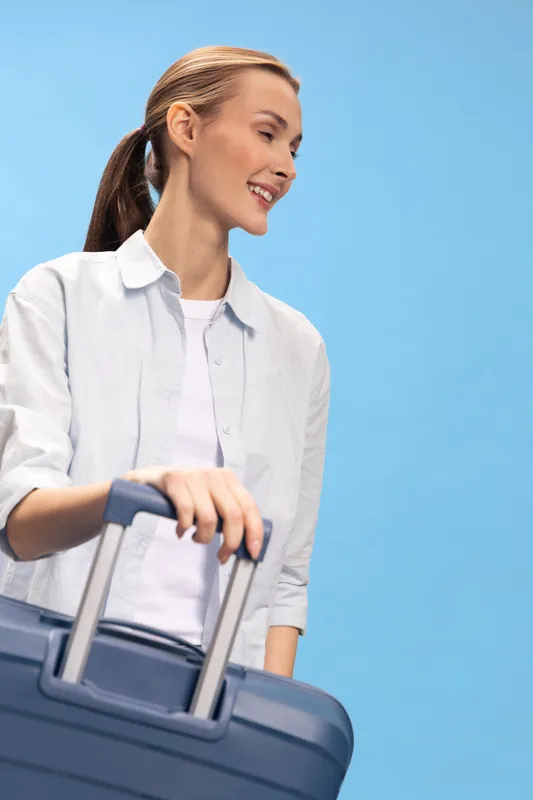 Smiling woman with ponytail holds suitcase handle, looking away on blue background. Concept for travel, vacation, and lifestyle with happy female traveler in casual clothes.