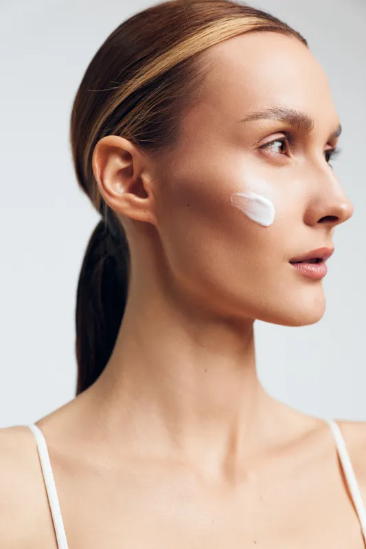A young woman applies cream to her cheek in a clean studio, with soft even lighting highlighting her focused, calm expression during a skincare routine.