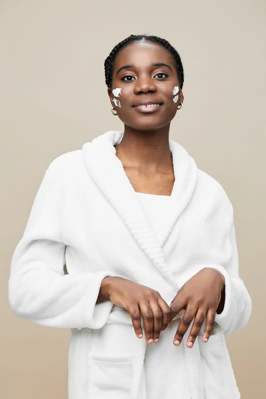 A young Black woman with braids smiles confidently while wearing a white bathrobe, showcasing skincare products on her face against a soft beige background