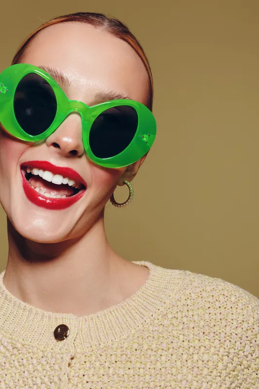 Smiling woman with bright red lipstick wearing large green sunglasses and a beige knitted sweater. Close-up portrait on a neutral background. Concept of fashion, style, and joyful mood.