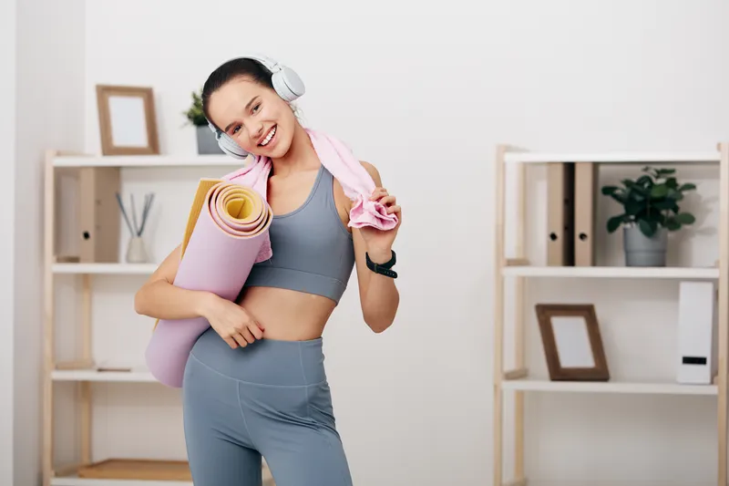 Fitness woman in athletic wear with yoga mat and headphones in a bright, minimalist room. She smiles, towel over shoulder, ready for home workout and fitness routine amid neat shelves and plants.