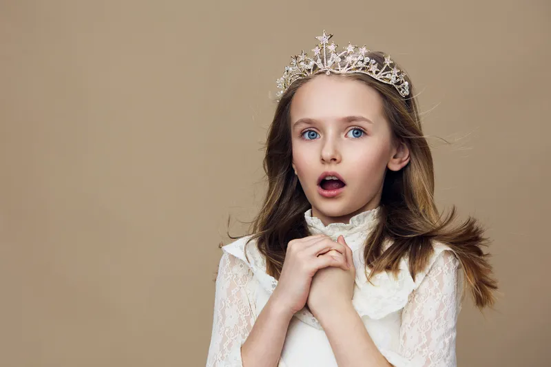 Surprised little girl in a white dress with a tiara, expressing wonder and excitement against a soft beige background