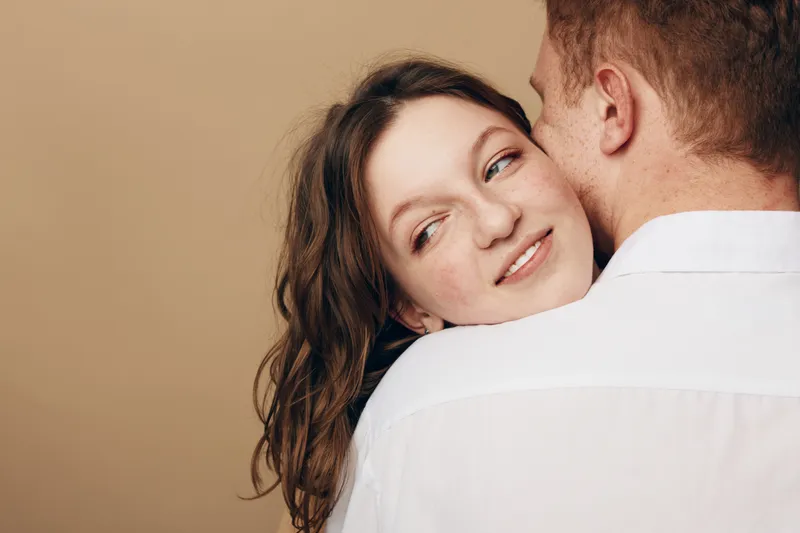 Young woman with brown hair smiles gently while embracing a man in white shirt against beige background. Concept of love, intimacy, trust, and close relationship between couple.