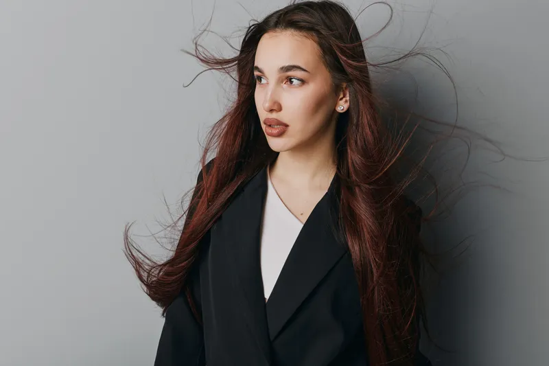 Young woman with long brown hair wearing black blazer and white top looking sideways on gray background. Studio portrait with natural makeup and loose hair in motion for fashion or beauty.