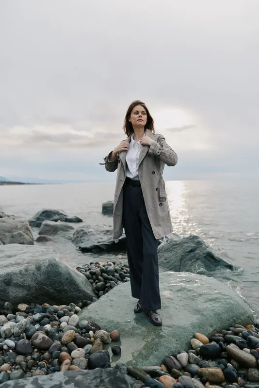 Woman in long coat stands on rocky shore near calm sea under cloudy sky. She looks confident and contemplative with hands adjusting coat collar. Concept of autumn fashion and nature.