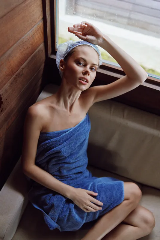 young woman wrapped in blue towel with shower cap sitting by wooden window, natural light highlighting relaxed expression and clean skincare routine indoors