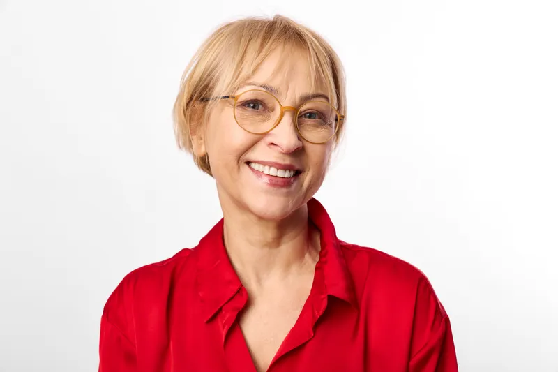 Happy mature woman portrait, smiling with round eyeglasses, short blond hair, wearing red shirt, looking at camera, soft studio light on clean minimal white background.