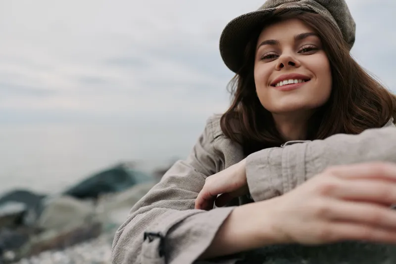 Smiling young woman in casual clothes and hat leans on a rock at the beach. Natural light highlights her relaxed mood and outdoor lifestyle on a cloudy day by the sea.