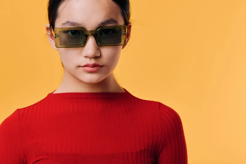 Woman wearing brown sunglasses and red shirt posing against yellow background looking forward with neutral expression for fashion or style portrait photography.