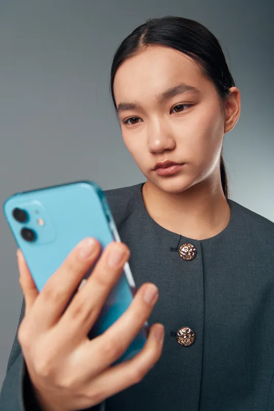 Young woman with dark hair wearing black coat looking at smartphone screen with focus and slight concern on gray background in closeup vertical portrait.