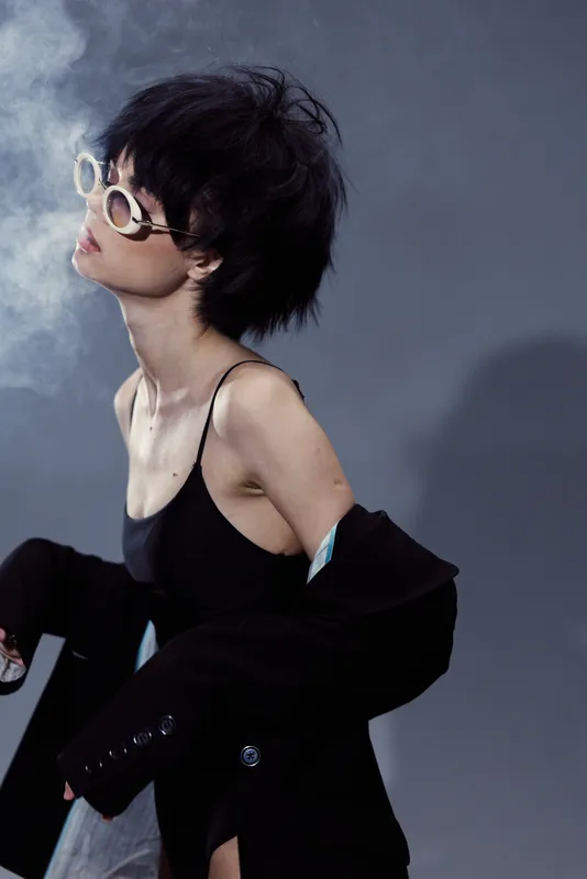 Stylish woman with short hair wearing sunglasses and black outfit, exhaling smoke in a moody studio setting.