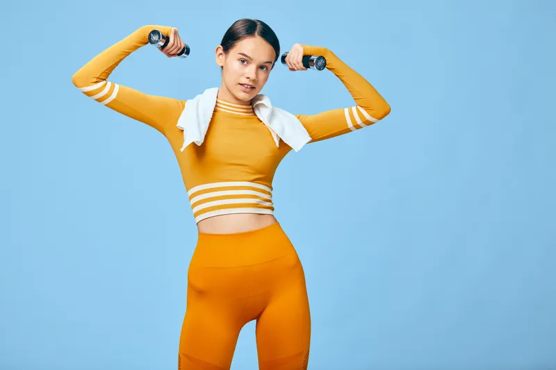 Woman in bright orange athleticwear flexes arms with a towel over her shoulders, standing in a blue studio. A dynamic fitness portrait showcasing strength, energy, and athletic confidence.