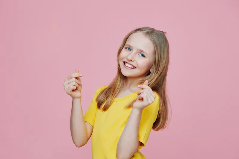 Happy young girl with long hair in a yellow t shirt smiling against a pink background, celebrating joy and positivity with playful gestures