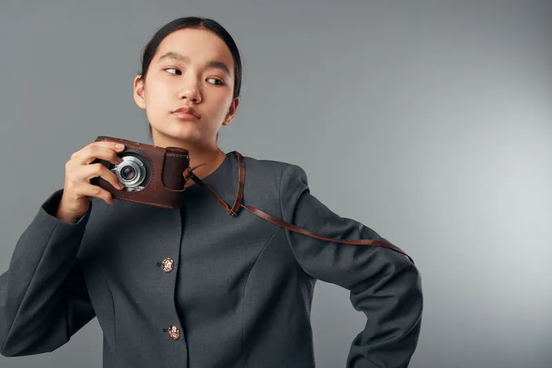 Young woman holding vintage camera in leather case, looking aside with serious expression. Fashion portrait with minimal background and casual jacket in studio lighting.