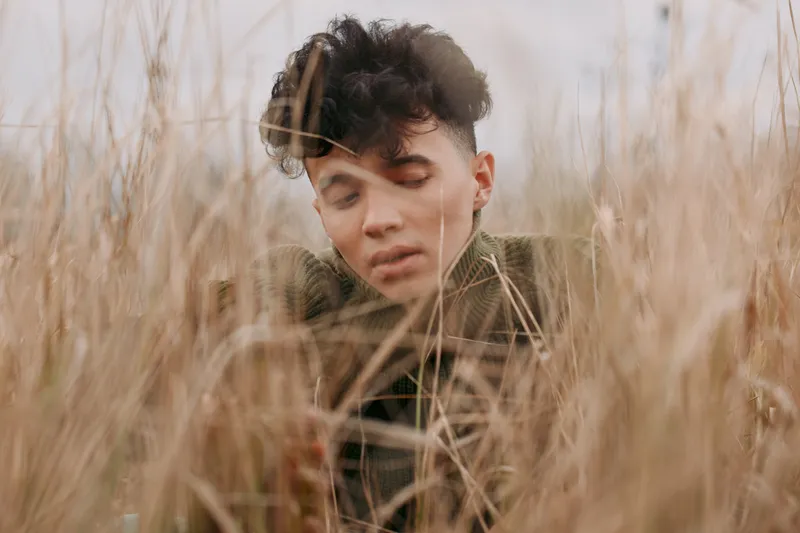 Close candid portrait of a young man nestled in tall dry grass, conveying genuine emotion and quiet presence; the image emphasizes authenticity and veracity through soft natural light and unposed
