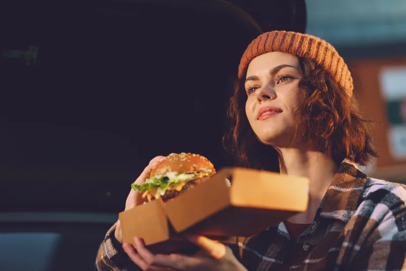 Woman holding burger in takeaway box by car at evening, portrait shot capturing authenticity and candid lifestyle with golden hour glow, mindful living, emotional storytelling, cozy beanie.