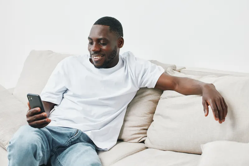 Happy young African man smiling while using smartphone on a cozy couch at home, highlighting modern lifestyle and technology