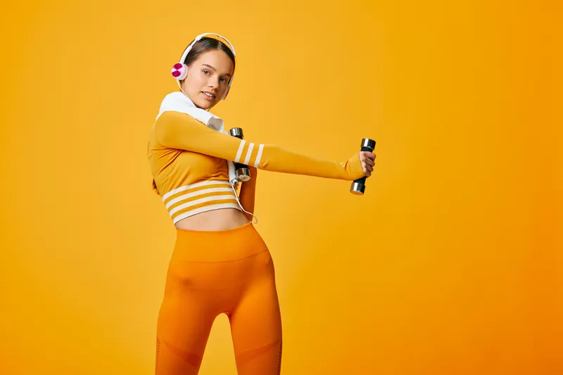 Woman fitness portrait showing sport workout with dumbbells and headphones in vibrant orange studio; activewear details, confident pose, toned core and minimal seamless background.